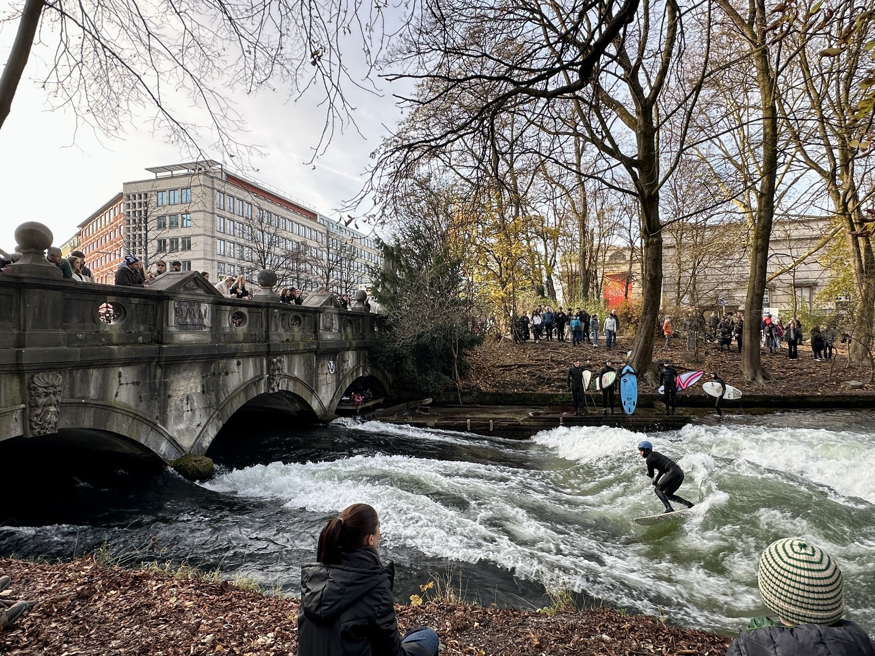 Surfer riding the Eisbach Wave in Munich's Englischer Garten, crowd watching from the bridge