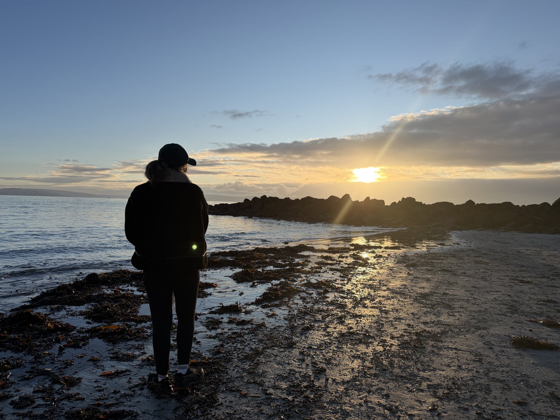 A person silhouetted against the golden sunset over Galway Bay, standing on the rocky shoreline