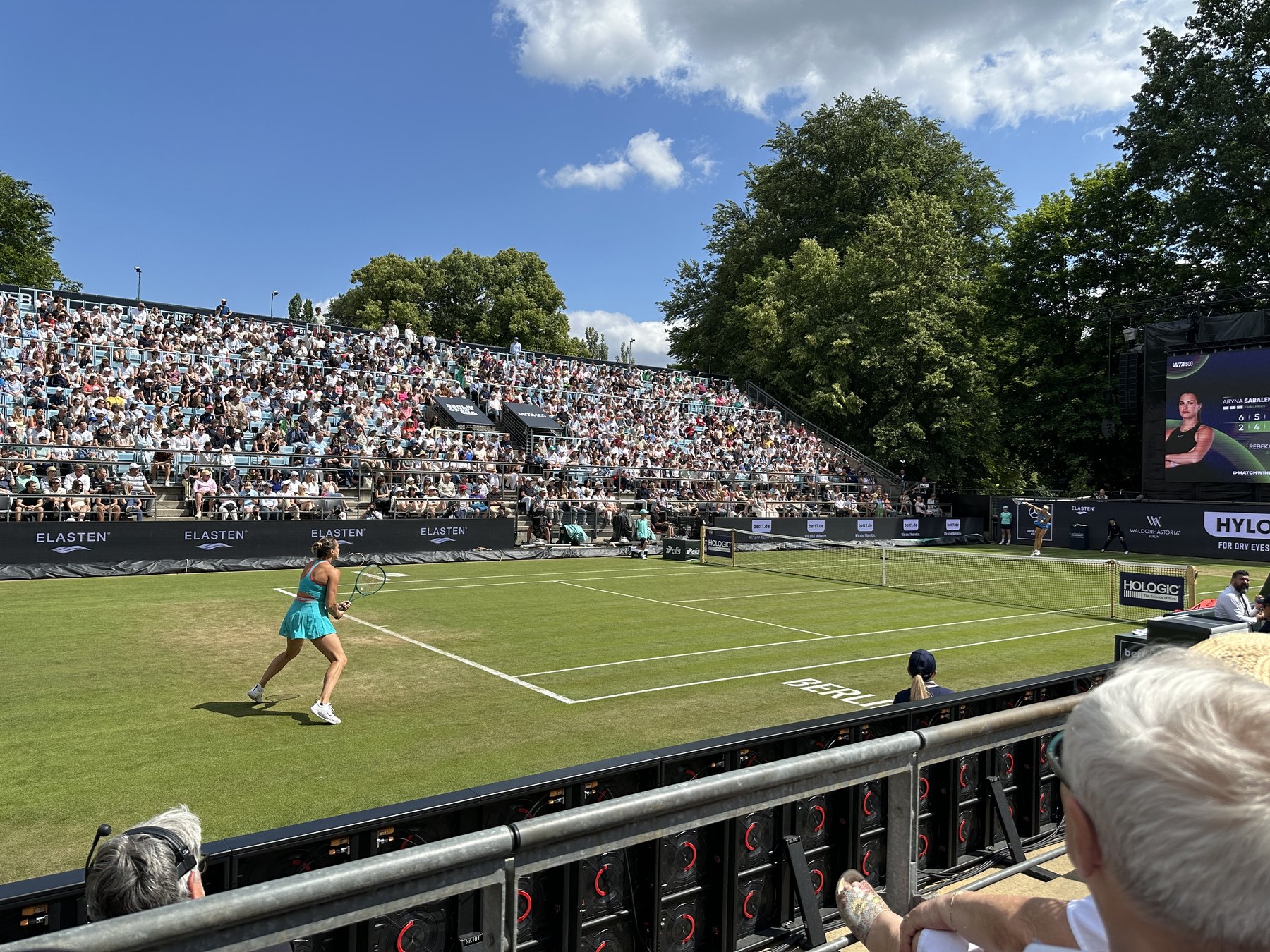 Aryna Sabalenka mid-swing on the grass court at the Berlin Open 2025, packed stands and blue summer sky behind her